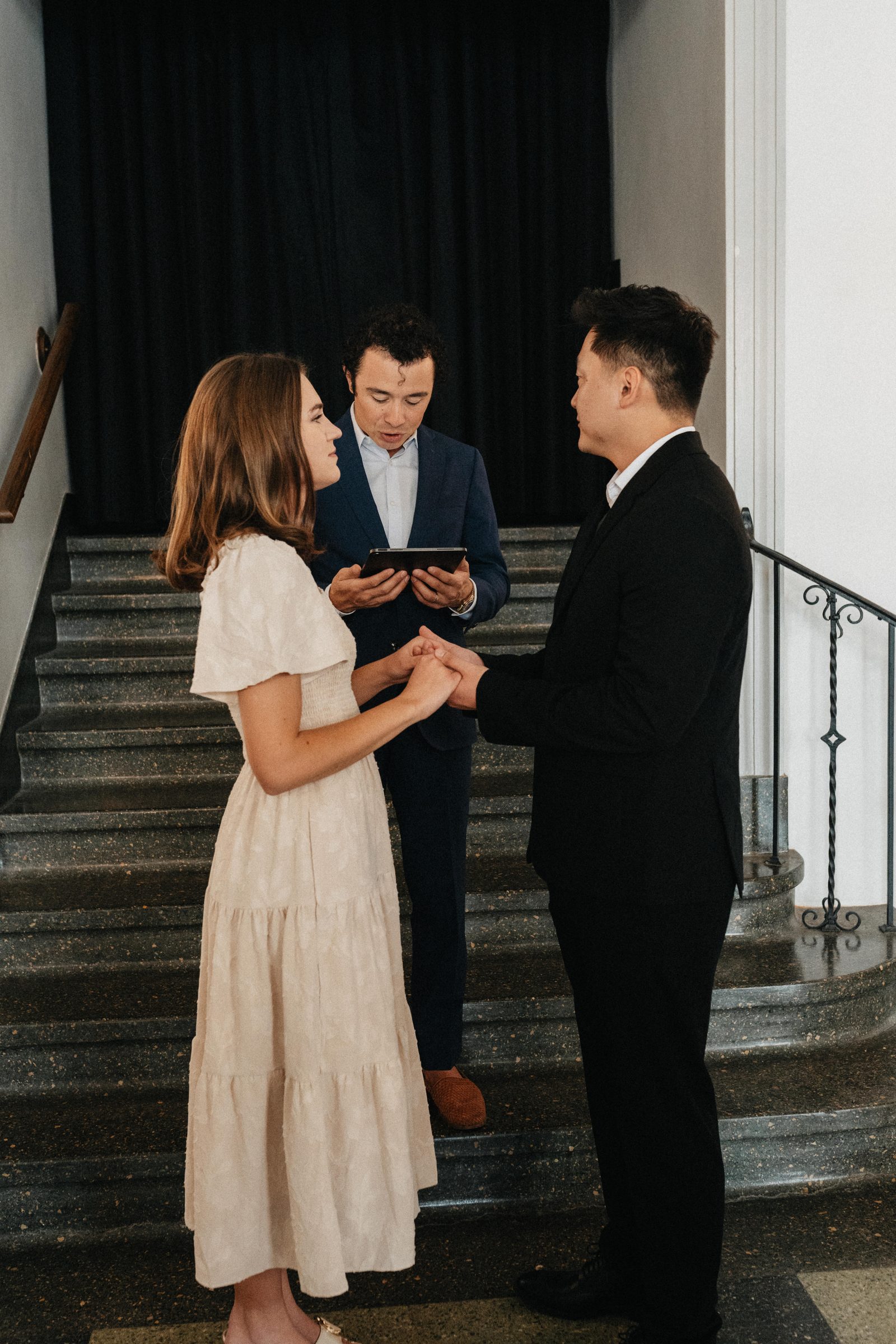 Couple holds hands on a grand staircase while the officiant reads during an intimate ceremony