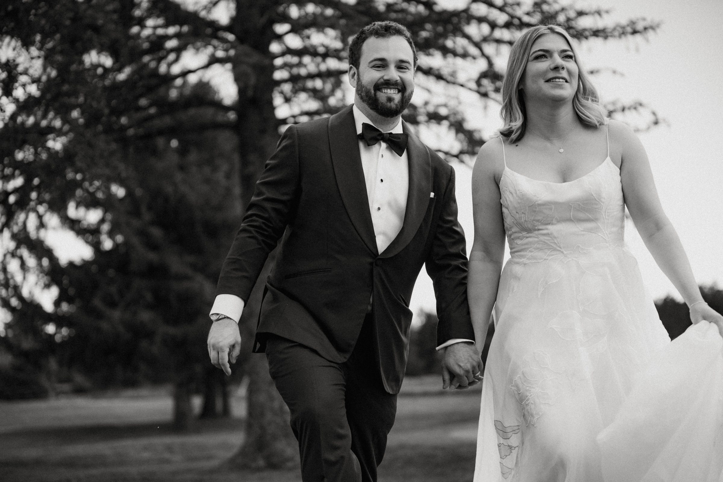 Black and white photo of a groom in a tuxedo and bride holding hands and walking together