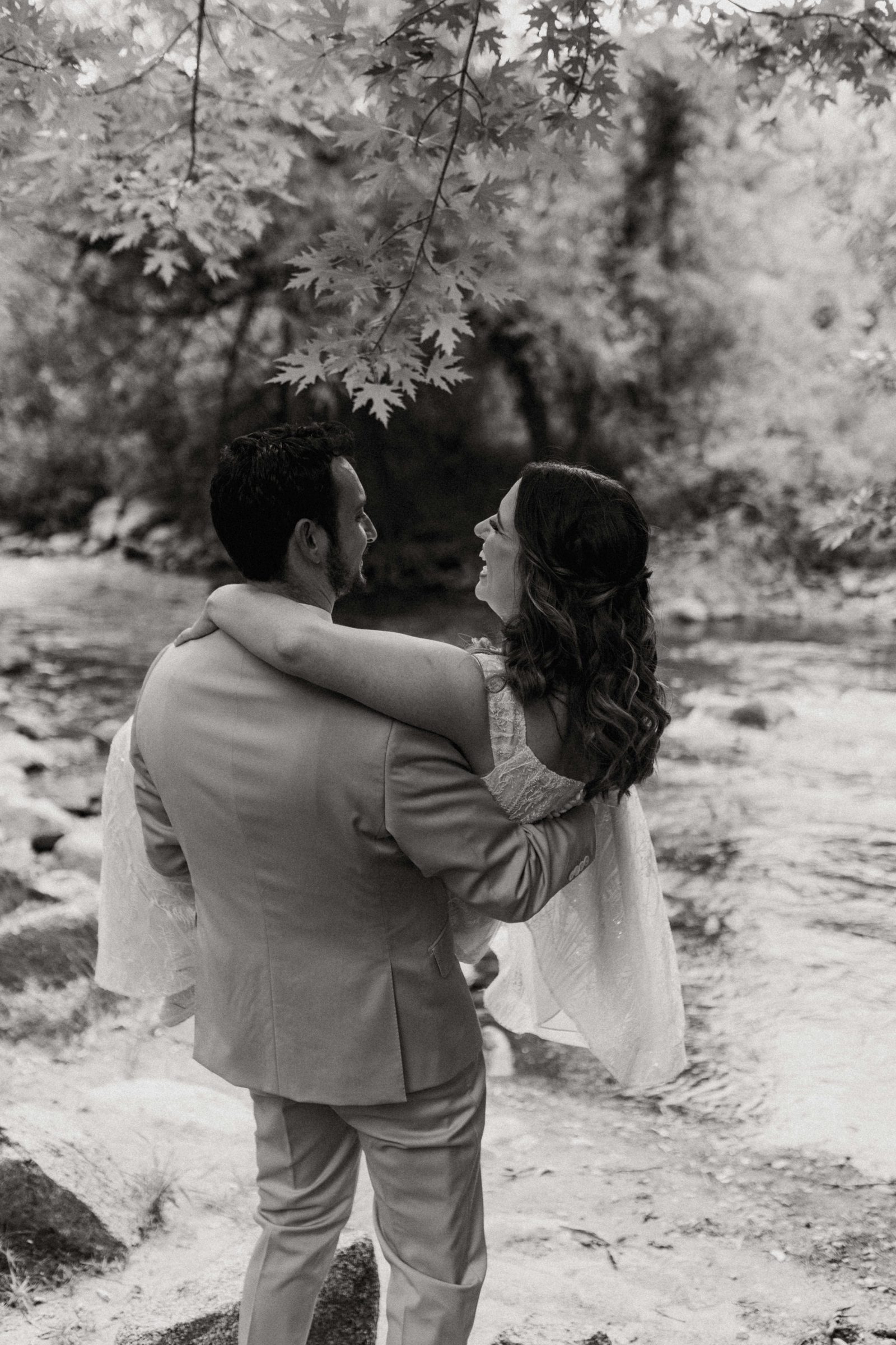 Black and white photo of a groom lifting his bride in an embrace beside a creek