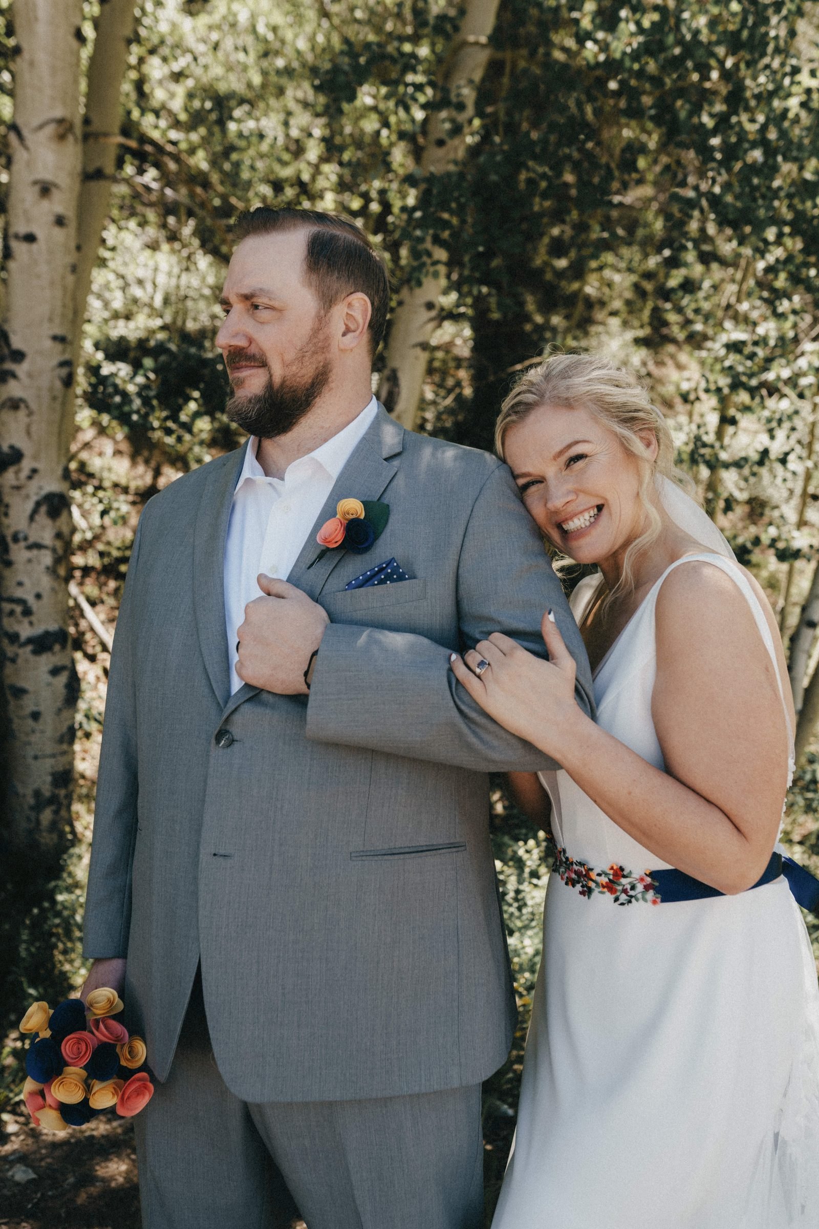Groom in a gray suit with his smiling bride tucked under his arm among aspen trees