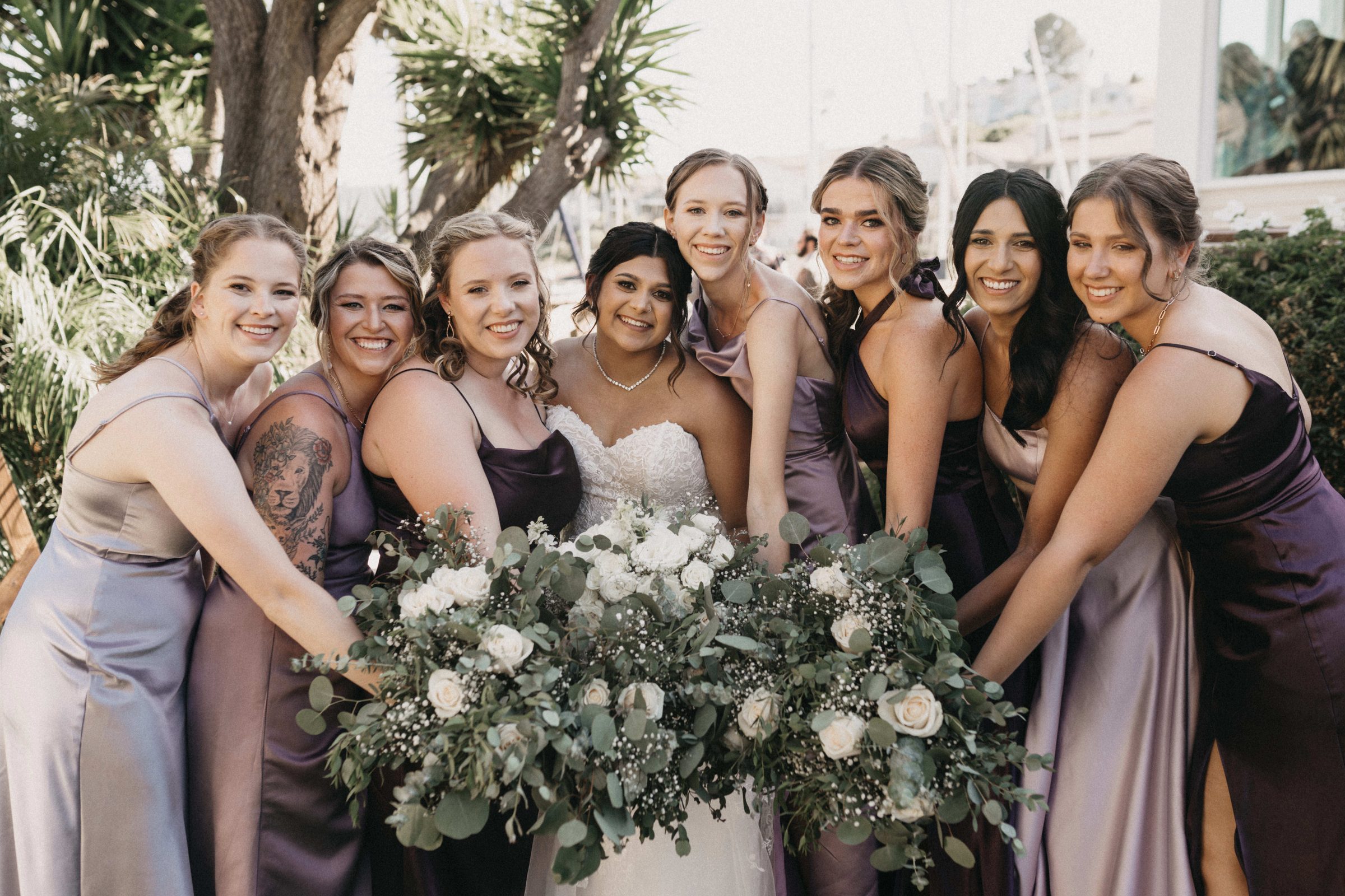 Bride and eight bridesmaids in lavender dresses laughing around a large bouquet