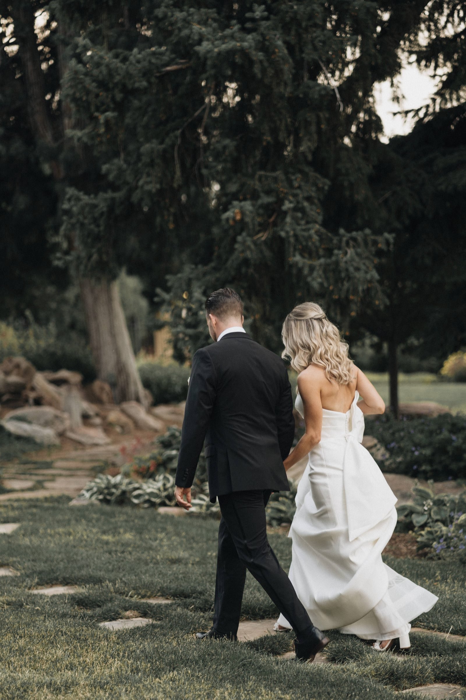 Groom and bride walking away from the camera along a stepping-stone path through a garden