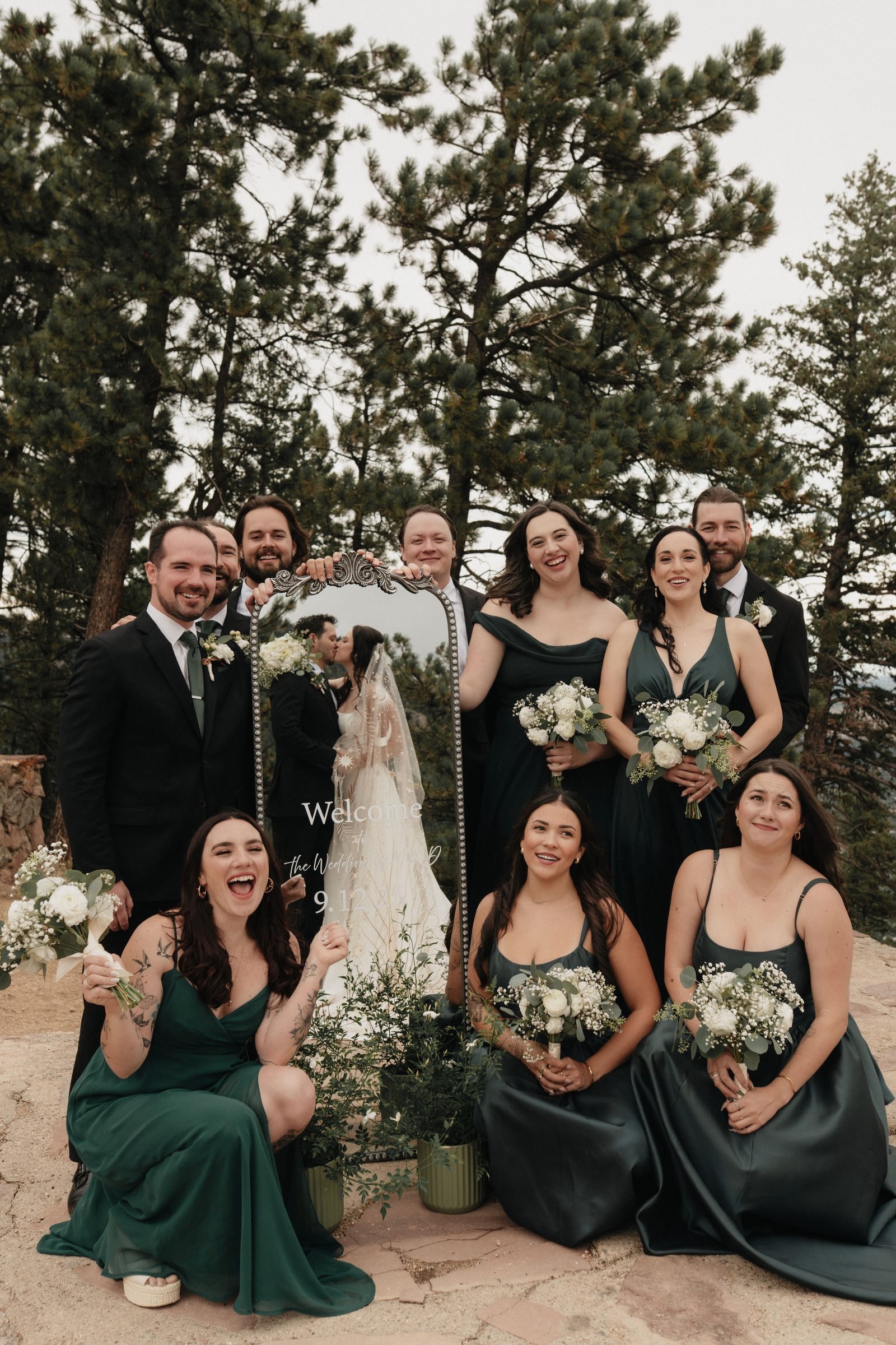Bridal party in emerald green dresses laughing together among pine trees with a mirror sign