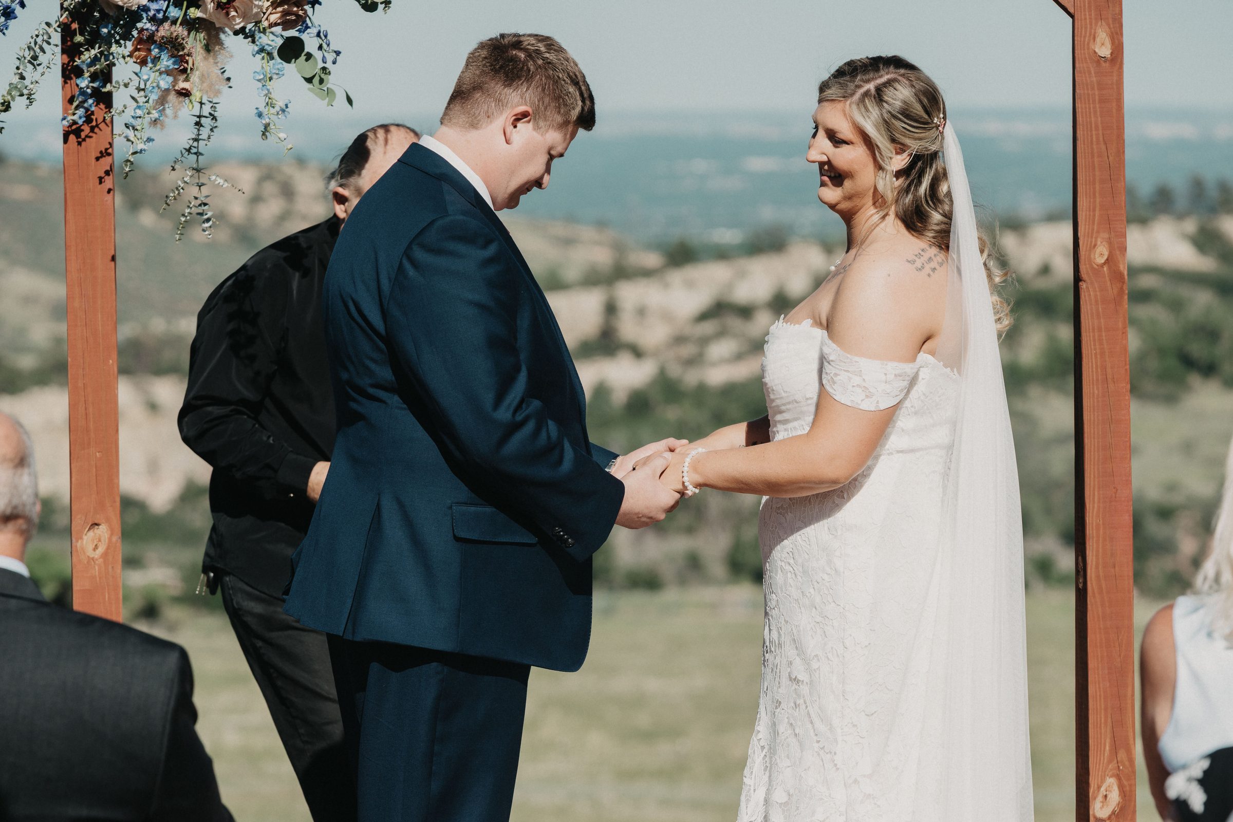 Bride and groom smile and hold hands under a rustic wooden arch above rolling hills
