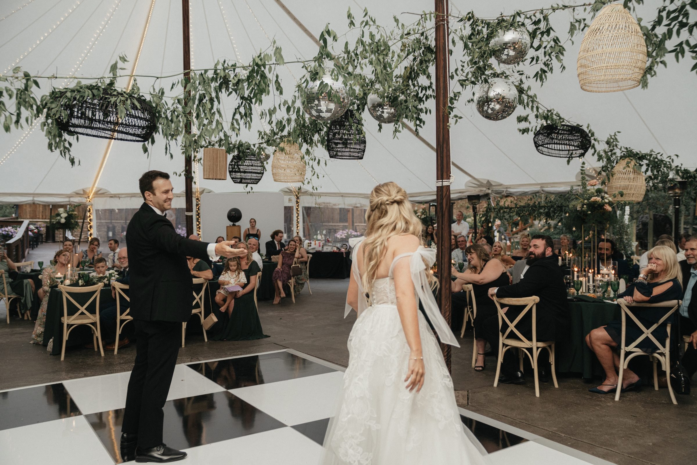 Bride and groom share a dance under a tented reception with hanging greenery and a checkerboard floor