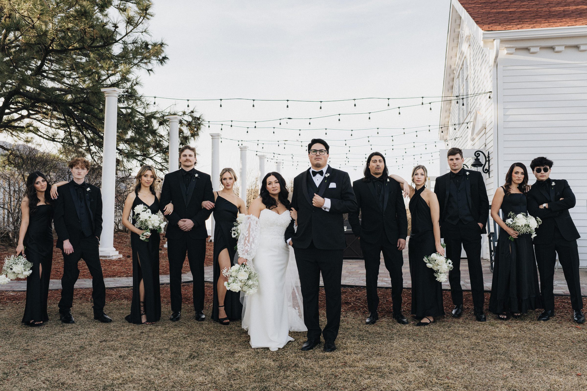 Wedding party in black standing in a line outside a white building with string lights