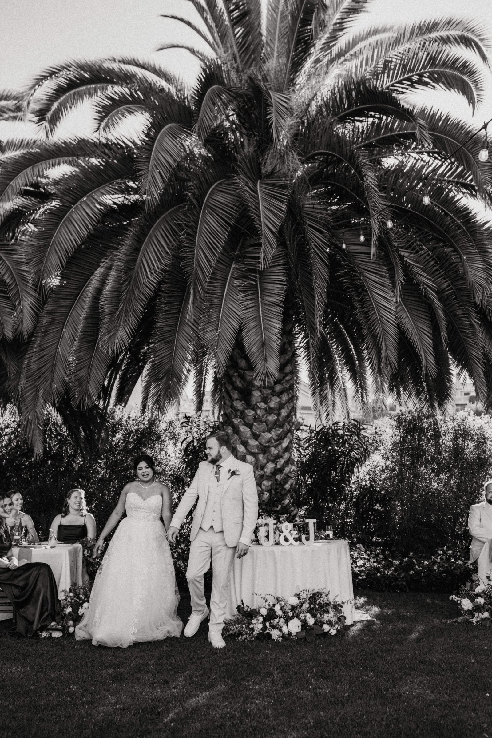 Black and white photo of a bride and groom walking to their sweetheart table beneath a palm tree