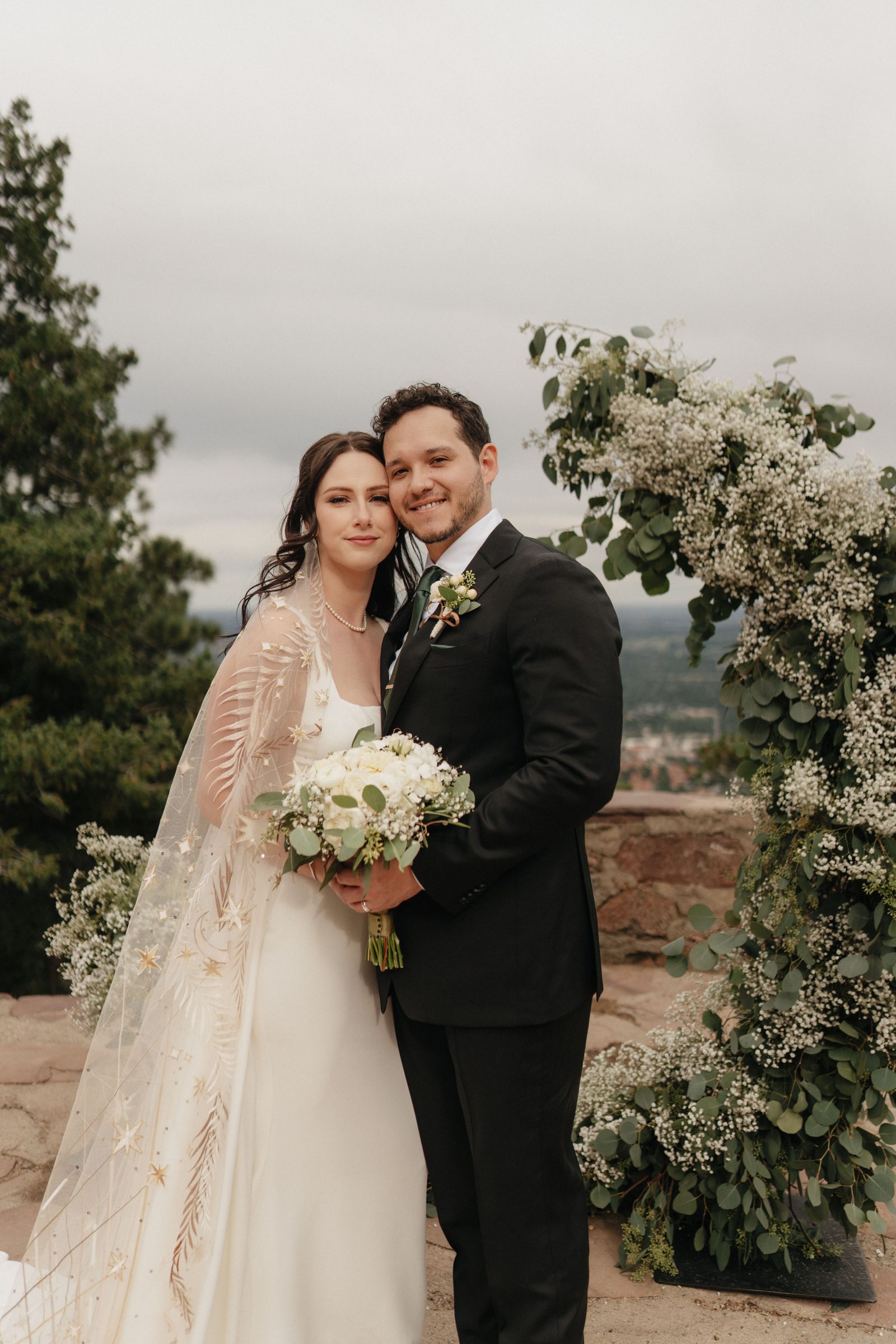 Bride and groom posing together beside a white floral arch at a hilltop ceremony site