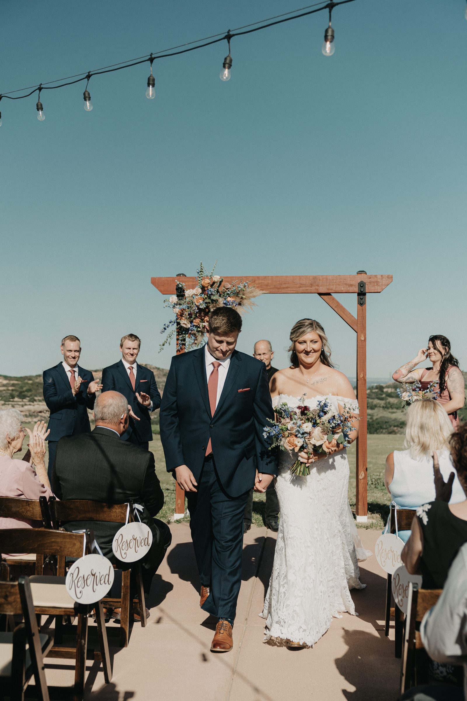 Newlyweds walk up the aisle after the ceremony under string lights as guests applaud