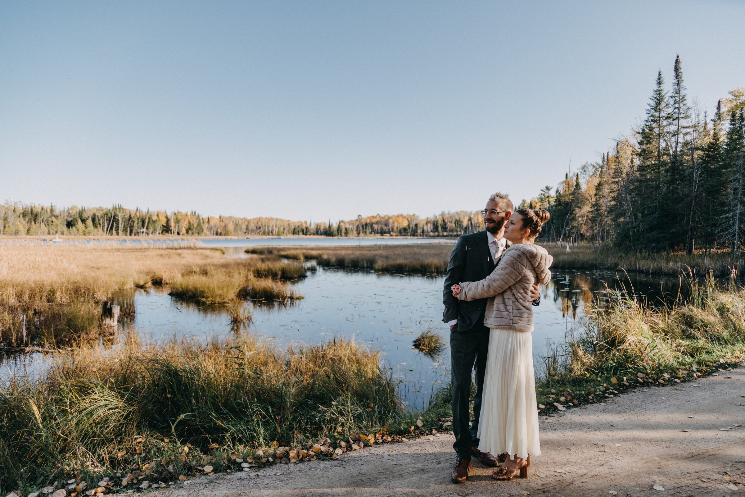 Bride in a fur wrap and groom in a dark suit embrace beside a marshy lake edged with evergreens and fall color