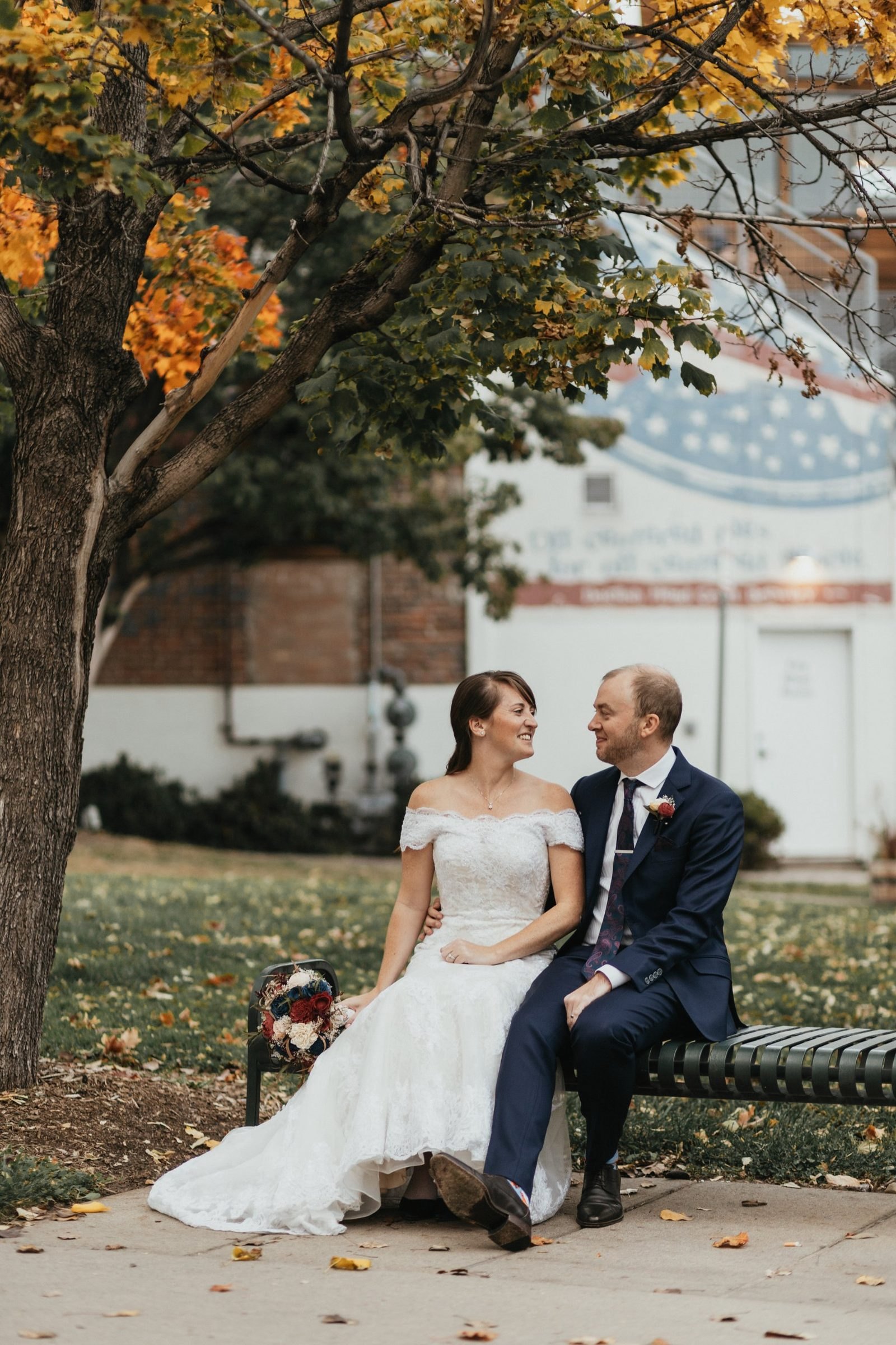 Bride and groom share a smiling look on a park bench beneath a maple tree in fall color